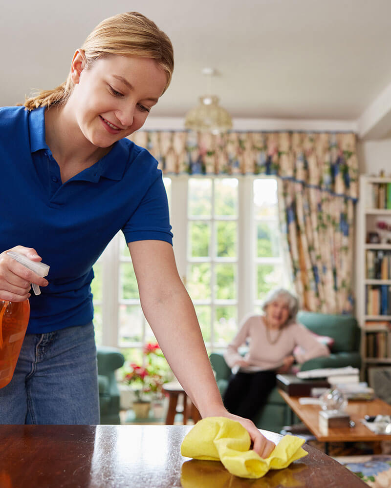 Young woman cleaning a table in the foreground with an elderly woman reading on a sofa chair in the background