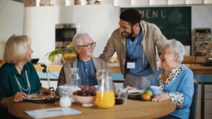 A male care worker wearing a lanyard and casual clothing standing and engaging warmly with three elderly residents seated around a wooden dining table during mealtime in a communal dining area with a menu board visible on the wall
