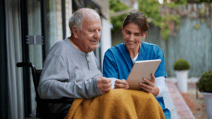 A healthcare worker in blue scrubs showing a tablet to an elderly man in a wheelchair covered with a yellow blanket, both smiling while looking at the screen together in a bright, modern care facility with glass walls and greenery in the background
