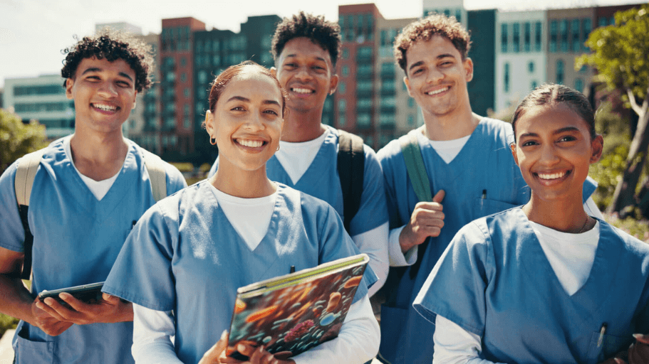 Group of smiling healthcare students in blue scrubs standing outdoors with backpacks and notebooks.