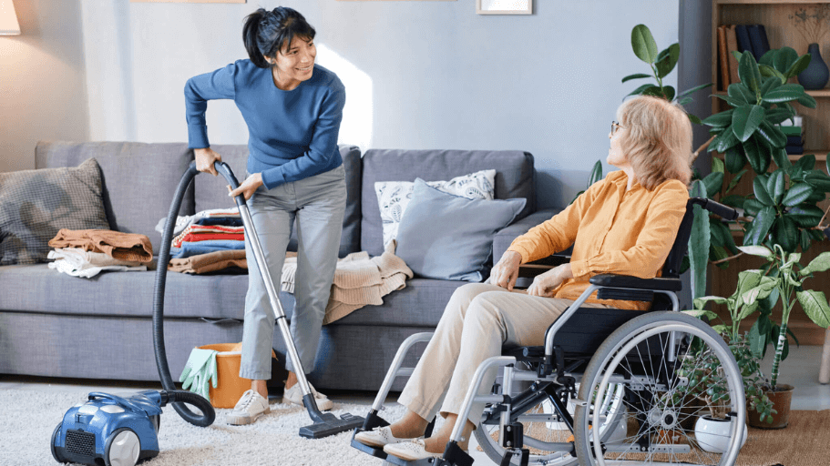 Caregiver vacuuming a living room while an older woman sits nearby in a wheelchair, watching and smiling.