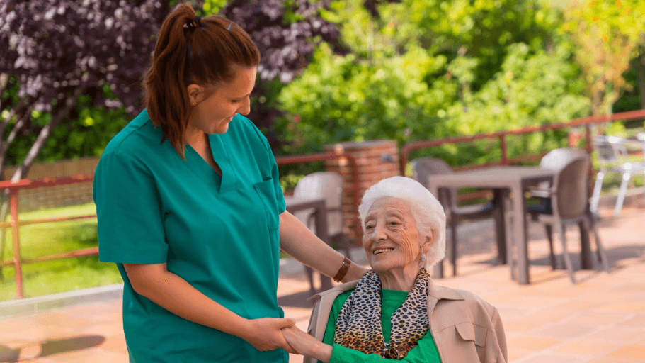 Caregiver in green scrubs walking beside an older woman seated outdoors on a patio, both smiling and holding hands.