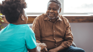 An older man and a younger person sitting together indoors, smiling and holding hands during a conversation.