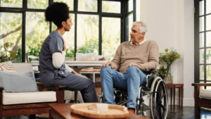 A caregiver sits with a man in a wheelchair in a bright, comfortable living space, engaged in conversation.