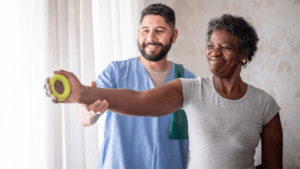 A caregiver guides an older woman through a light arm exercise with a handheld weight, both smiling in a bright indoor setting.