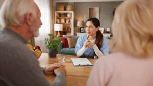 A caregiver meets with an older couple at a table in a comfortable home setting, discussing care and support services.